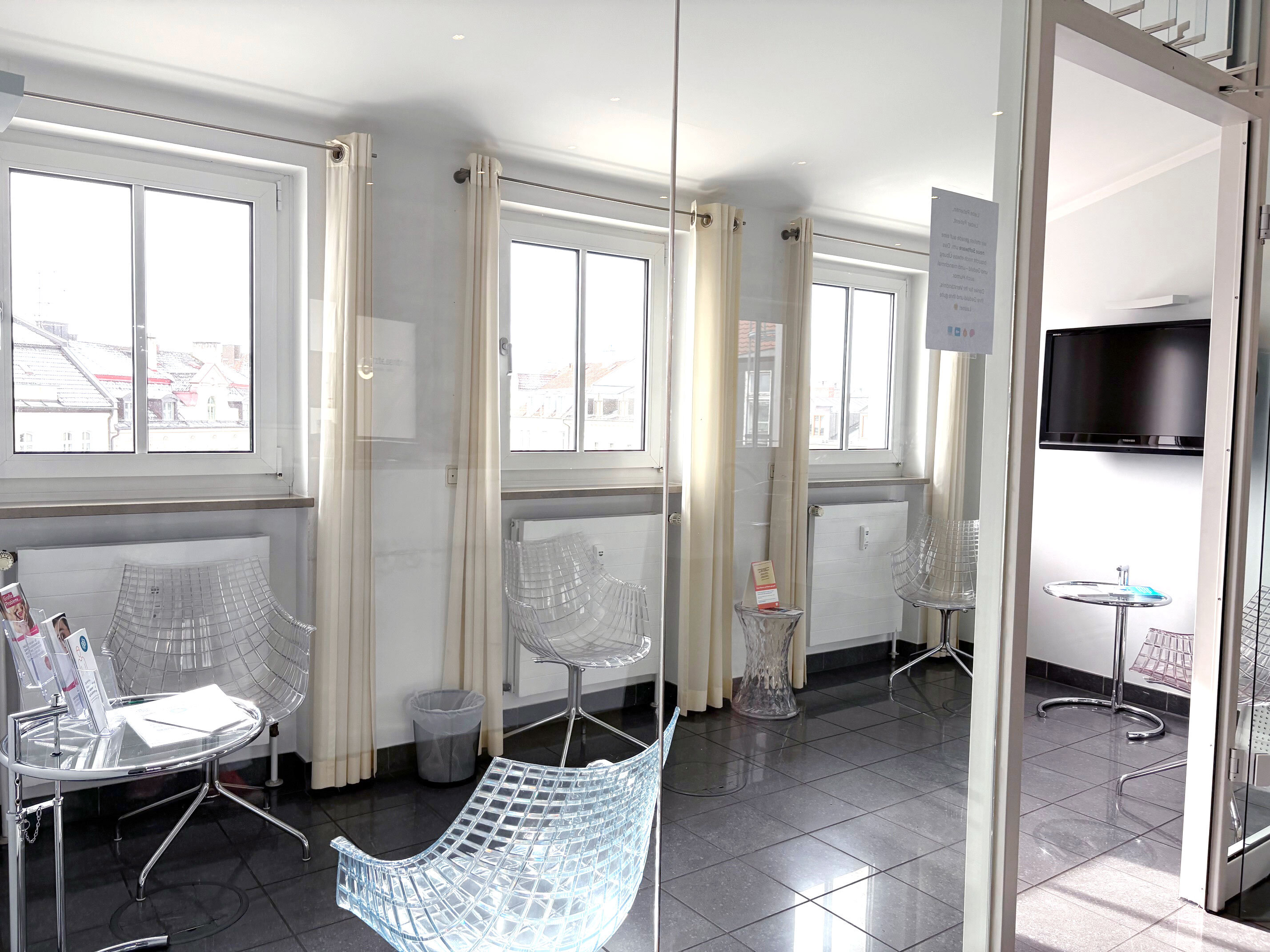 Modernes, helles Wartezimmer mit großen Fenstern im Ärztezentrum Einsteinstraße, transparenten Stühlen und Glastischen mit Infomaterial.