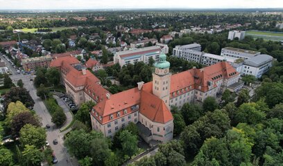 Luftaufnahme eines historischen Gebäudes mit Uhrenturm, umgeben von Bäumen und weiteren Stadtgebäuden.
