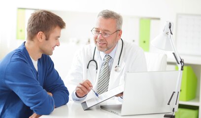 Allgemeinmediziner mit Stethoskop berät jungen Patienten am Schreibtisch in moderner Praxis, beide im Gespräch. 
